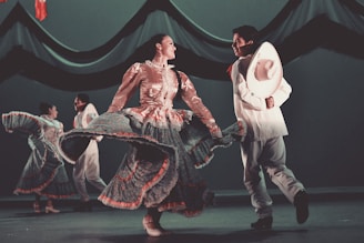 A group of dancers performing in traditional attire, showcasing colorful and flowing dresses with intricate patterns and a man holding a white hat. The setting is a stage with dark drapery in the background.