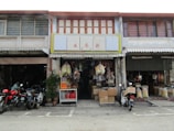 A traditional shopfront with a variety of goods displayed, including bags of produce, boxes, and hanging items. Motorcycles are parked in front on a paved area. The building has a rustic look with wooden elements and signs with Chinese characters.