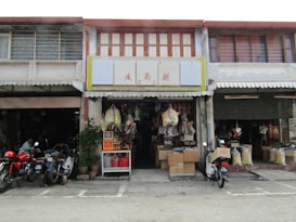 A traditional shopfront with a variety of goods displayed, including bags of produce, boxes, and hanging items. Motorcycles are parked in front on a paved area. The building has a rustic look with wooden elements and signs with Chinese characters.