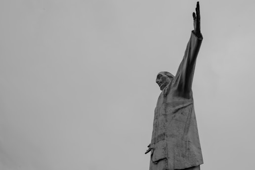 A towering statue of a person with one arm raised against a gray, overcast sky. The figure is clothed in a suit and gazes upward, conveying a sense of authority and grandeur.