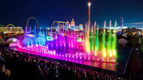 A vibrant nighttime display with colorful fountains illuminated in shades of pink, blue, and green, accompanied by a large audience in the foreground. Amusement park rides and bright lights can be seen in the background, providing a festive and lively atmosphere.
