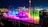 A vibrant nighttime display with colorful fountains illuminated in shades of pink, blue, and green, accompanied by a large audience in the foreground. Amusement park rides and bright lights can be seen in the background, providing a festive and lively atmosphere.