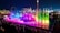 A vibrant nighttime display with colorful fountains illuminated in shades of pink, blue, and green, accompanied by a large audience in the foreground. Amusement park rides and bright lights can be seen in the background, providing a festive and lively atmosphere.