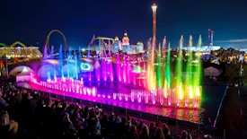A vibrant nighttime display with colorful fountains illuminated in shades of pink, blue, and green, accompanied by a large audience in the foreground. Amusement park rides and bright lights can be seen in the background, providing a festive and lively atmosphere.