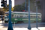 A vintage streetcar painted in green and cream travels through an urban area, partially obscured by a blue traffic pole. The streetcar has signage indicating destinations like Balboa Park and San Diego Zoo. Several pedestrians are visible near the intersections, and the street is lined with trees and urban infrastructure.