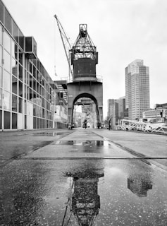 A large industrial crane is positioned along a paved walkway by the waterfront. The crane's reflection is visible in a puddle on the ground. Surrounding the area are modern high-rise buildings, with cloudy skies overhead, creating a monochrome and industrial atmosphere.