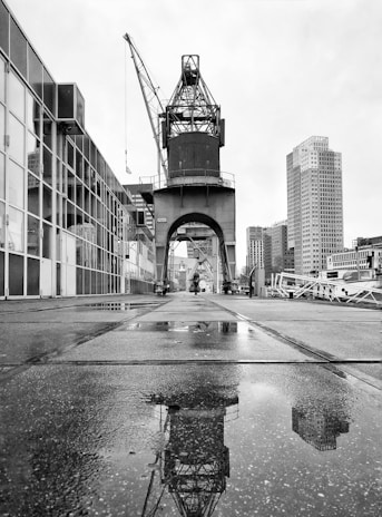 A large industrial crane is positioned along a paved walkway by the waterfront. The crane's reflection is visible in a puddle on the ground. Surrounding the area are modern high-rise buildings, with cloudy skies overhead, creating a monochrome and industrial atmosphere.