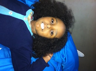 A child lies on two blue blankets or cushions, wearing a navy blue cardigan with light blue edges. The child's hair is thick and curly, and a piece of candy or a small object is in their mouth.