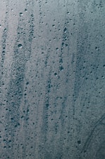 Condensation with water droplets is visible on a glass surface. The droplets vary in size and are densely packed, creating an intricate pattern. The light reflects off the droplets, giving a glistening effect.