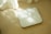 A digital scale with a sleek, square design lies on a beige tiled floor. Sunlight filters through sheer curtains, casting gentle shadows and soft light patterns on the floor and the surface of the scale.