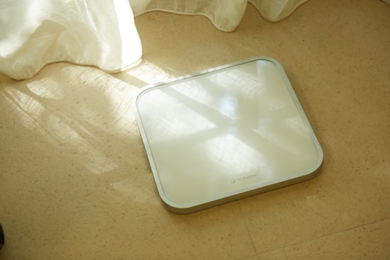 A digital scale with a sleek, square design lies on a beige tiled floor. Sunlight filters through sheer curtains, casting gentle shadows and soft light patterns on the floor and the surface of the scale.