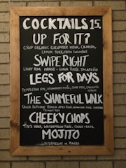 A chalkboard with a wooden frame lists various cocktails and their ingredients. The drinks include 'Up For It?' with cucumber vodka and cilantro, 'Swipe Right' with rum and guava purée, 'Legs For Days' with rye and strawberry purée, 'The Shameful Walk' with tequila and apple purée, 'Cheeky Chops' with vodka and watermelon purée, and a 'Mojito' with either watermelon or mango.