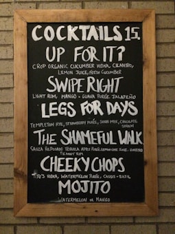 A chalkboard with a wooden frame lists various cocktails and their ingredients. The drinks include 'Up For It?' with cucumber vodka and cilantro, 'Swipe Right' with rum and guava purée, 'Legs For Days' with rye and strawberry purée, 'The Shameful Walk' with tequila and apple purée, 'Cheeky Chops' with vodka and watermelon purée, and a 'Mojito' with either watermelon or mango.
