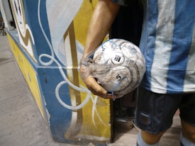 A person wearing a blue and white striped jersey is holding a soccer ball wrapped in plastic. The scene is set against a vibrant mural with swirling patterns in blue, yellow, and white. The setting appears to be outdoors, possibly near a street or alley.