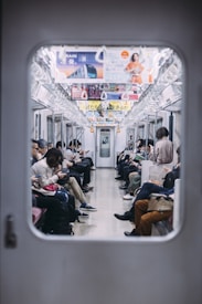A crowded train carriage with passengers seated on either side, many of whom are engaged with their smartphones. Overhead, various advertisements can be seen attached to the train's interior. The scene conveys the routine of daily commuting.