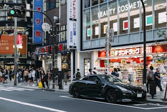 A busy urban street scene featuring a crowd of people walking near a store with bright signage. A sleek black sports car is prominently parked in front. Various advertisements and shop signs fill the background, contributing to the bustling city atmosphere.