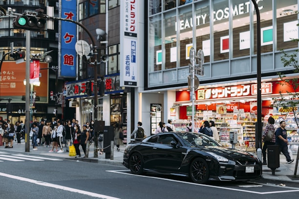 A busy urban street scene featuring a crowd of people walking near a store with bright signage. A sleek black sports car is prominently parked in front. Various advertisements and shop signs fill the background, contributing to the bustling city atmosphere.