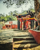 A small outdoor temple structure adorned with colorful fabric is visible, surrounded by a variety of trees and greenery. The temple is constructed with wooden beams and red painted walls, and there seem to be steps leading up to it. Another small structure with red walls and a board with text in a local language is visible in the background.