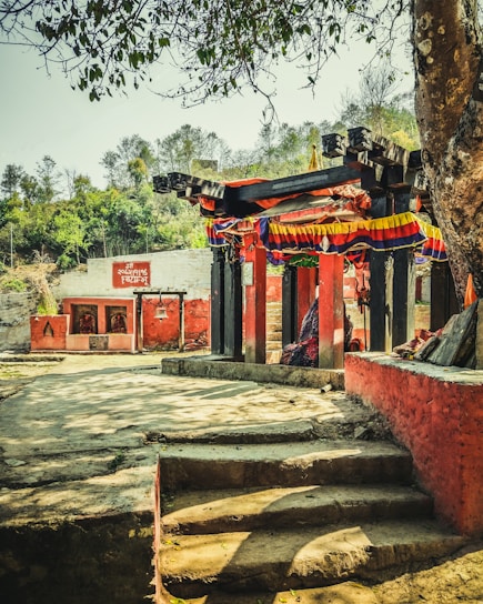 A small outdoor temple structure adorned with colorful fabric is visible, surrounded by a variety of trees and greenery. The temple is constructed with wooden beams and red painted walls, and there seem to be steps leading up to it. Another small structure with red walls and a board with text in a local language is visible in the background.