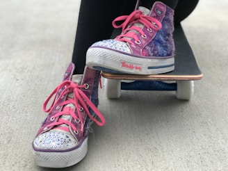 A pair of colorful high-top sneakers with glitter and rhinestones are positioned on a skateboard. The sneakers have pink shoelaces and a small heart logo on the side. The skateboard is on a concrete surface.