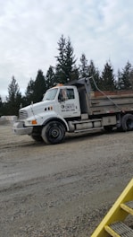 A large white construction truck is parked on a gravel area with trees in the background under an overcast sky.