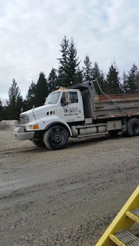 A large white construction truck is parked on a gravel area with trees in the background under an overcast sky.