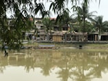 A serene riverside scene with traditional Vietnamese-style buildings in the background, surrounded by lush greenery and palm trees. The water in the foreground reflects the structures and foliage, creating a tranquil atmosphere. Bamboo leaves hang down from the top, adding to the tropical feel. Wooden boats are docked by the waterside, contributing to the rustic charm.