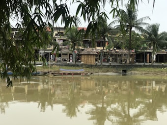 A serene riverside scene with traditional Vietnamese-style buildings in the background, surrounded by lush greenery and palm trees. The water in the foreground reflects the structures and foliage, creating a tranquil atmosphere. Bamboo leaves hang down from the top, adding to the tropical feel. Wooden boats are docked by the waterside, contributing to the rustic charm.