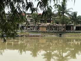 A serene riverside scene with traditional Vietnamese-style buildings in the background, surrounded by lush greenery and palm trees. The water in the foreground reflects the structures and foliage, creating a tranquil atmosphere. Bamboo leaves hang down from the top, adding to the tropical feel. Wooden boats are docked by the waterside, contributing to the rustic charm.