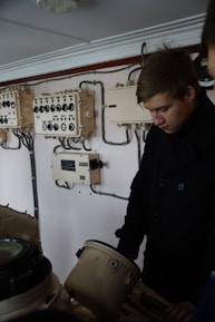 A person is standing near an array of vintage electrical control panels. The walls are equipped with various switches and dials connected by wires. The individual appears to be wearing a dark coat and is looking downward at some equipment.