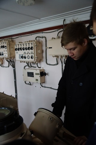 A person is standing near an array of vintage electrical control panels. The walls are equipped with various switches and dials connected by wires. The individual appears to be wearing a dark coat and is looking downward at some equipment.