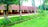 A long, single-story building with a red-tiled roof and white walls is surrounded by lush greenery. Coconut trees and a grassy lawn are in the foreground, and a dirt path runs parallel to the building. Wooden pillars are spaced evenly between the trees.