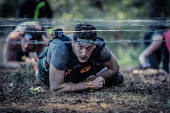 A focused participant in athletic gear is crawling under a barbed wire obstacle during an outdoor race or challenge. The surroundings include dirt and small plants, indicating a rugged environment. Other participants can be seen attempting the same challenge in the background.