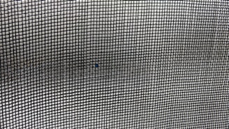 A close-up view of a mesh screen with a grid pattern. The material appears metallic with a series of small, evenly spaced square openings creating a repetitive and uniform pattern. The focus is on the grid, with slight variations in light and shadow visible across the surface.