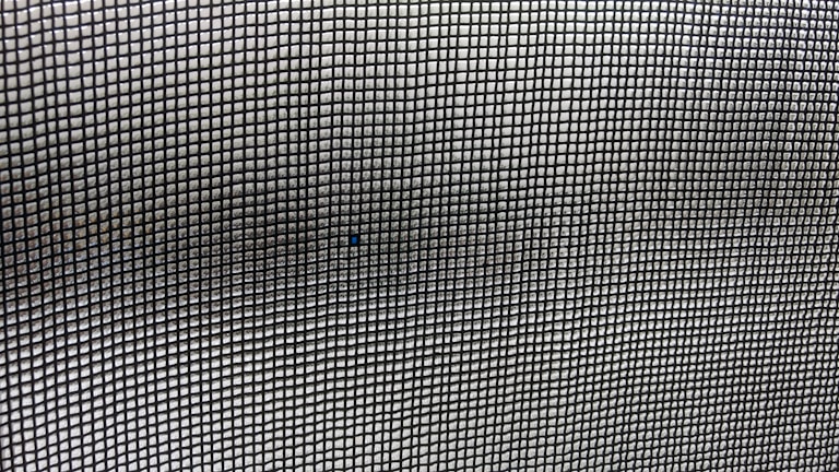 A close-up view of a mesh screen with a grid pattern. The material appears metallic with a series of small, evenly spaced square openings creating a repetitive and uniform pattern. The focus is on the grid, with slight variations in light and shadow visible across the surface.