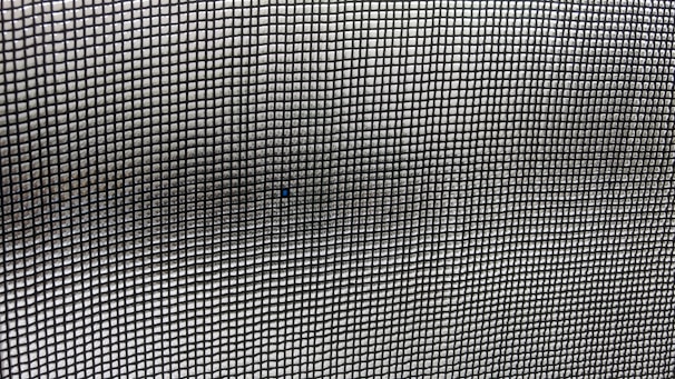 A close-up view of a mesh screen with a grid pattern. The material appears metallic with a series of small, evenly spaced square openings creating a repetitive and uniform pattern. The focus is on the grid, with slight variations in light and shadow visible across the surface.