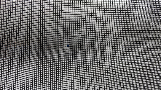 A close-up view of a mesh screen with a grid pattern. The material appears metallic with a series of small, evenly spaced square openings creating a repetitive and uniform pattern. The focus is on the grid, with slight variations in light and shadow visible across the surface.
