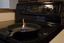 A black cast iron pan is placed on the surface of an electric stove. A small contained flame is burning in the center of the pan. The stove has multiple control knobs and a digital clock displaying 12:08. The surrounding area includes a wooden edge and a section of a white countertop.