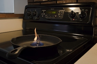A black cast iron pan is placed on the surface of an electric stove. A small contained flame is burning in the center of the pan. The stove has multiple control knobs and a digital clock displaying 12:08. The surrounding area includes a wooden edge and a section of a white countertop.
