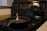 A black cast iron pan is placed on the surface of an electric stove. A small contained flame is burning in the center of the pan. The stove has multiple control knobs and a digital clock displaying 12:08. The surrounding area includes a wooden edge and a section of a white countertop.