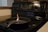 A black cast iron pan is placed on the surface of an electric stove. A small contained flame is burning in the center of the pan. The stove has multiple control knobs and a digital clock displaying 12:08. The surrounding area includes a wooden edge and a section of a white countertop.