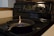 A black cast iron pan is placed on the surface of an electric stove. A small contained flame is burning in the center of the pan. The stove has multiple control knobs and a digital clock displaying 12:08. The surrounding area includes a wooden edge and a section of a white countertop.