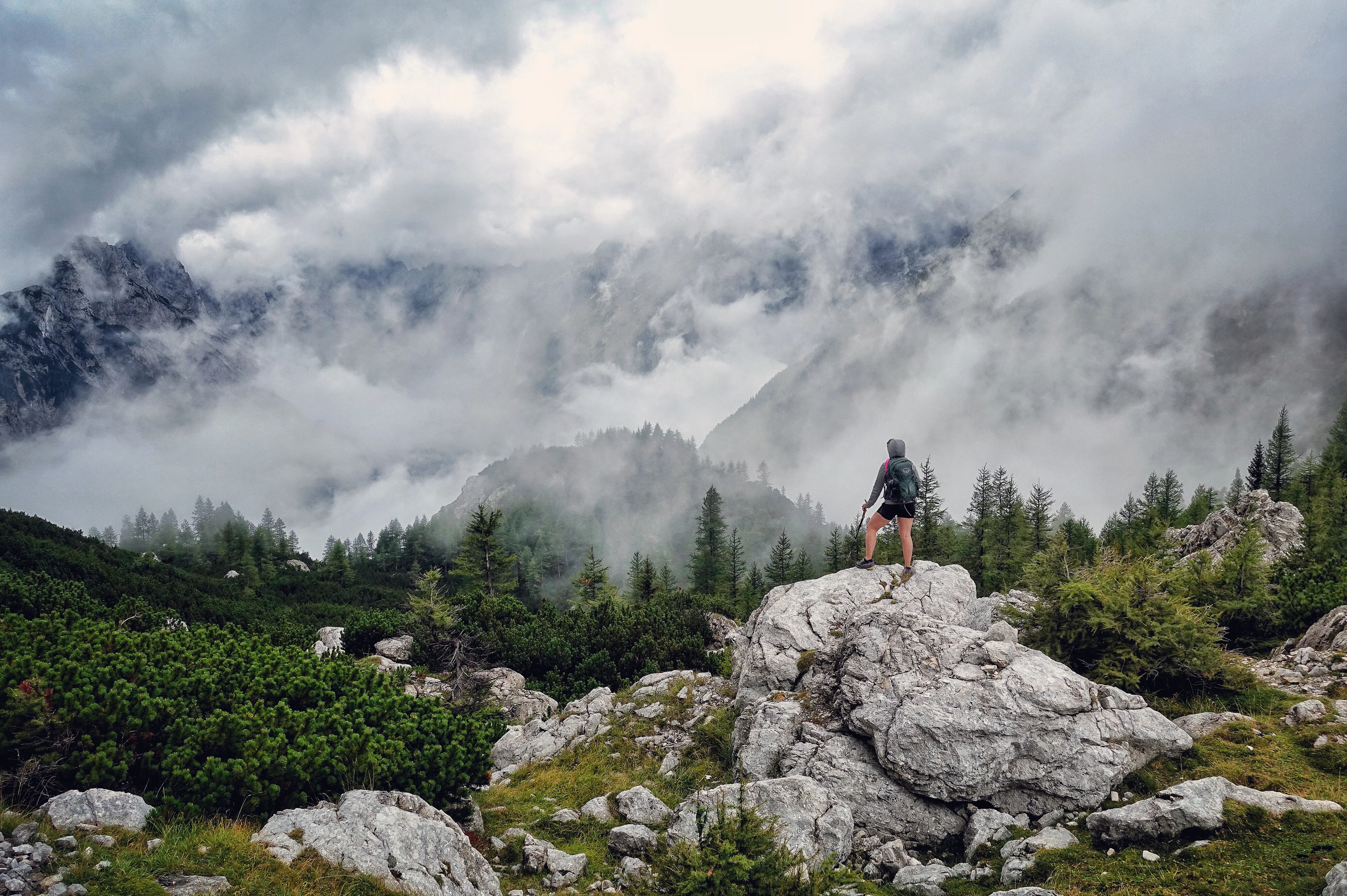 A traveller on a boulder looking out at cloud-wrapped limestone peaks on the Vršič Pass