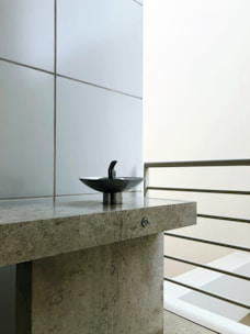 A modern sink with a sleek metal fixture is centered on a textured stone countertop. The background consists of large, smooth tiles and a set of metal railings, suggesting a contemporary design. The lighting is soft, emphasizing the minimalist aesthetic.