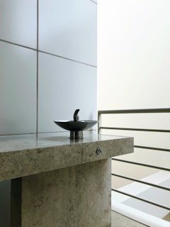 A modern sink with a sleek metal fixture is centered on a textured stone countertop. The background consists of large, smooth tiles and a set of metal railings, suggesting a contemporary design. The lighting is soft, emphasizing the minimalist aesthetic.