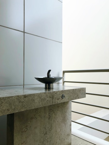 A modern sink with a sleek metal fixture is centered on a textured stone countertop. The background consists of large, smooth tiles and a set of metal railings, suggesting a contemporary design. The lighting is soft, emphasizing the minimalist aesthetic.