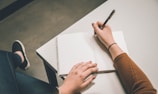A person is seated at a desk, writing in a blank notebook with a pen. The person is wearing a brown sweater and a wristband. The focus is on the hands and the notebook, with one hand resting on the page and the other holding the pen.