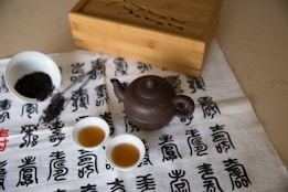 A traditional tea setup featuring a small dark teapot and two cups filled with tea. A bowl containing loose tea leaves is present alongside a metal scoop. The items are placed on a cloth adorned with calligraphy and a wooden box sits in the background.