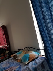 A bed featuring a colorful bedsheet and a pillow is positioned near a window with blue curtains. To the left, there is a red quilted item stacked against the wall, possibly a mattress or blanket. The room has a simple, minimalistic decor with a plain white wall.