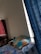 A bed featuring a colorful bedsheet and a pillow is positioned near a window with blue curtains. To the left, there is a red quilted item stacked against the wall, possibly a mattress or blanket. The room has a simple, minimalistic decor with a plain white wall.